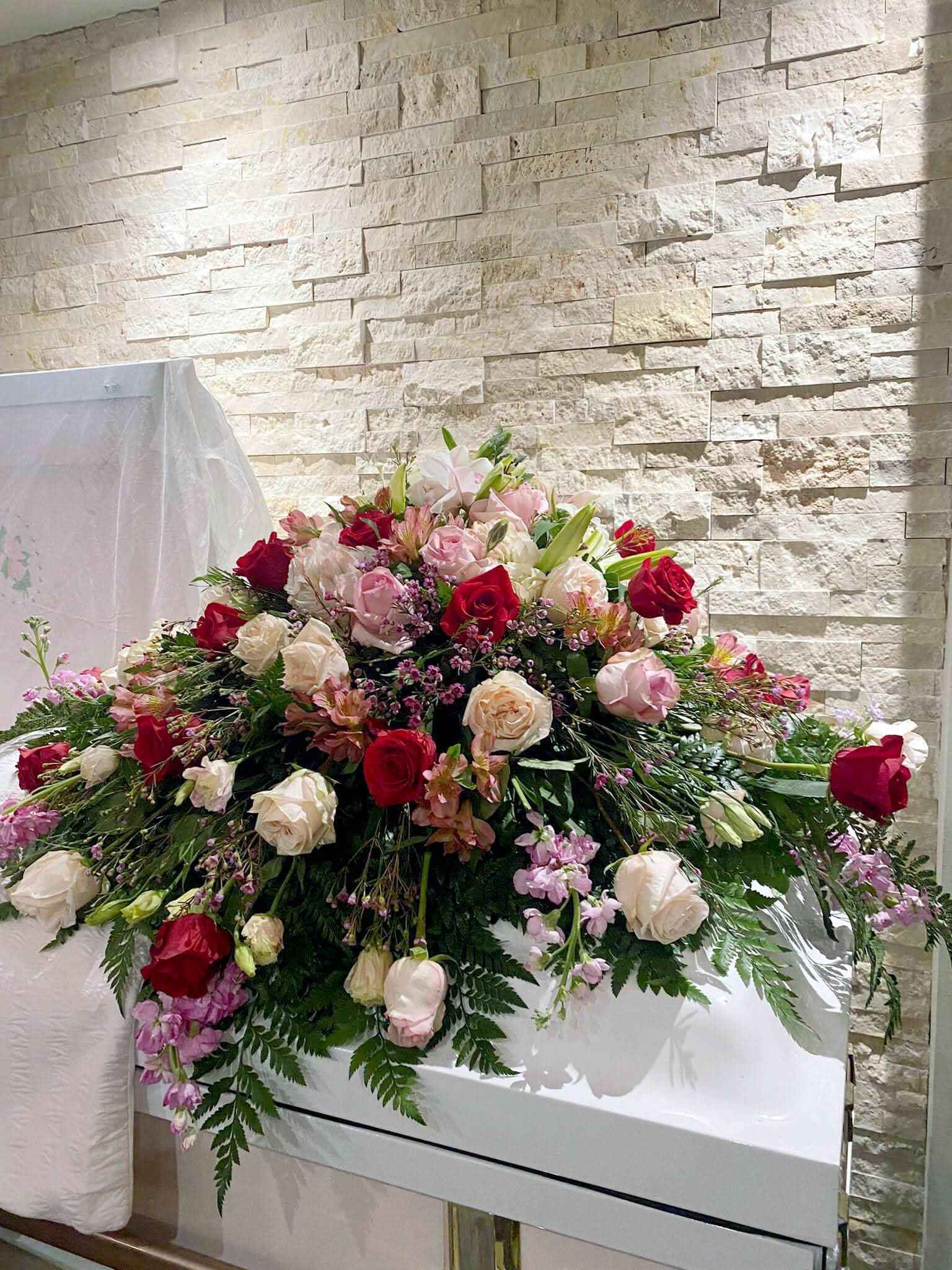 Arreglo floral funerario con rosas rojas y rosas sobre ataúd blanco, fondo de pared clara.