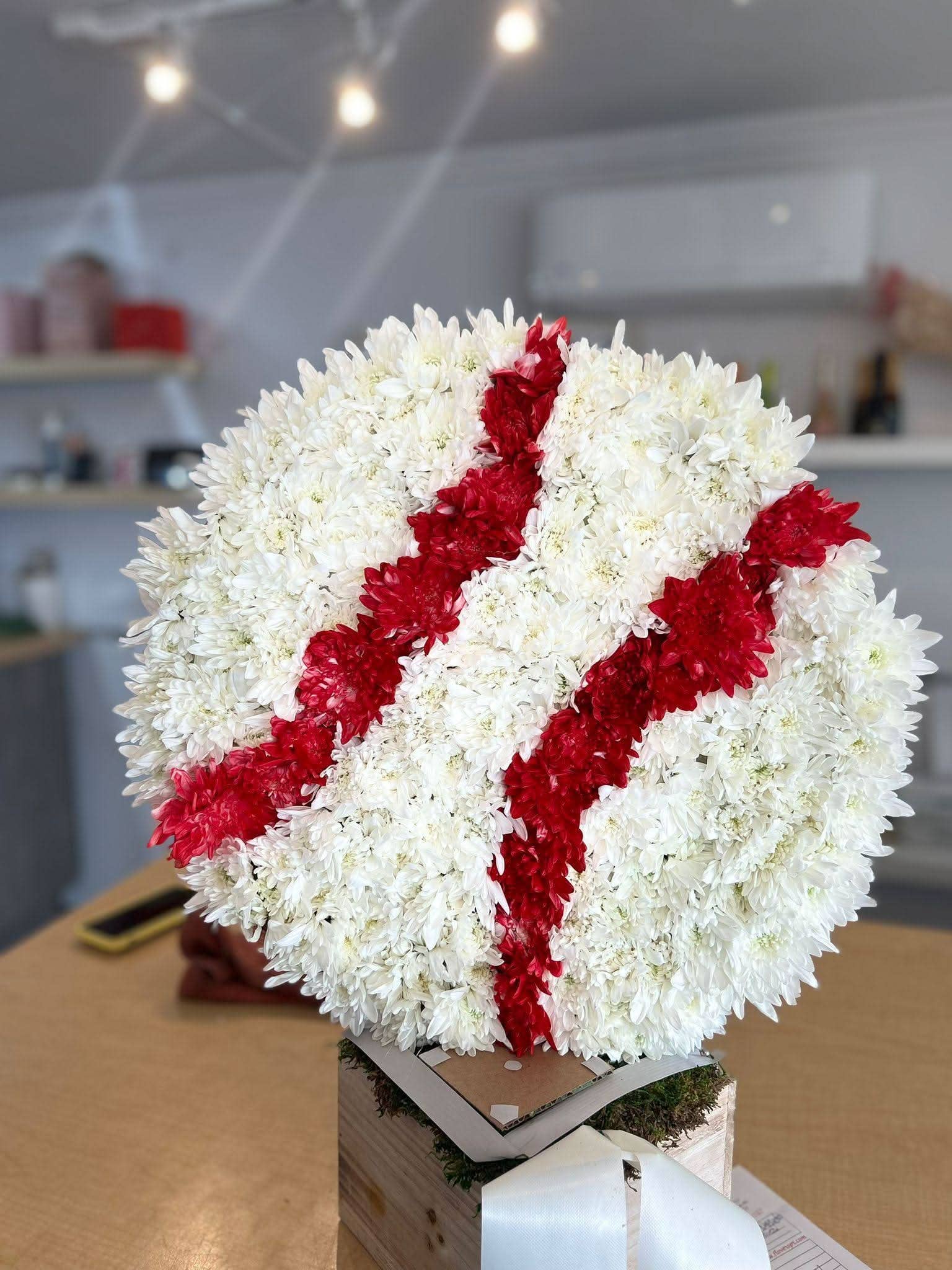 Arreglo floral redondo en forma de pelota de béisbol con flores blancas y detalles rojos, sobre base de madera.