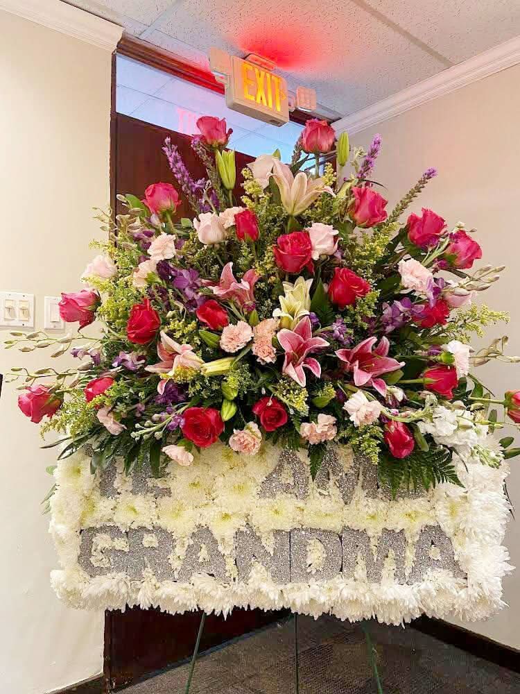 Corona funeraria de flores frescas con rosas rojas, lirios rosados y mensaje 'GRANDMA'.