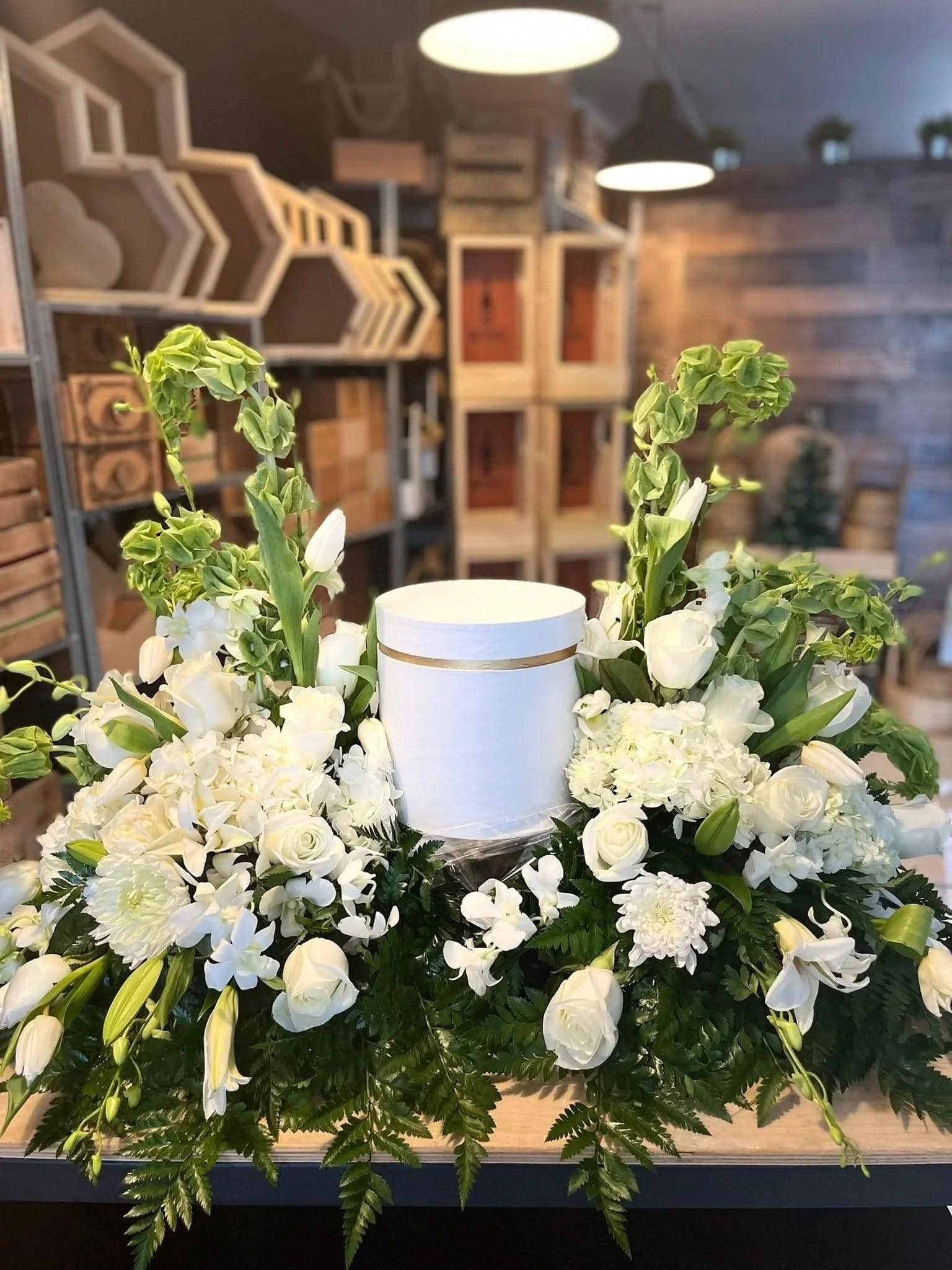 Arreglo floral funeral blanco con rosas, lirios y follaje, porta cenizas en centro.