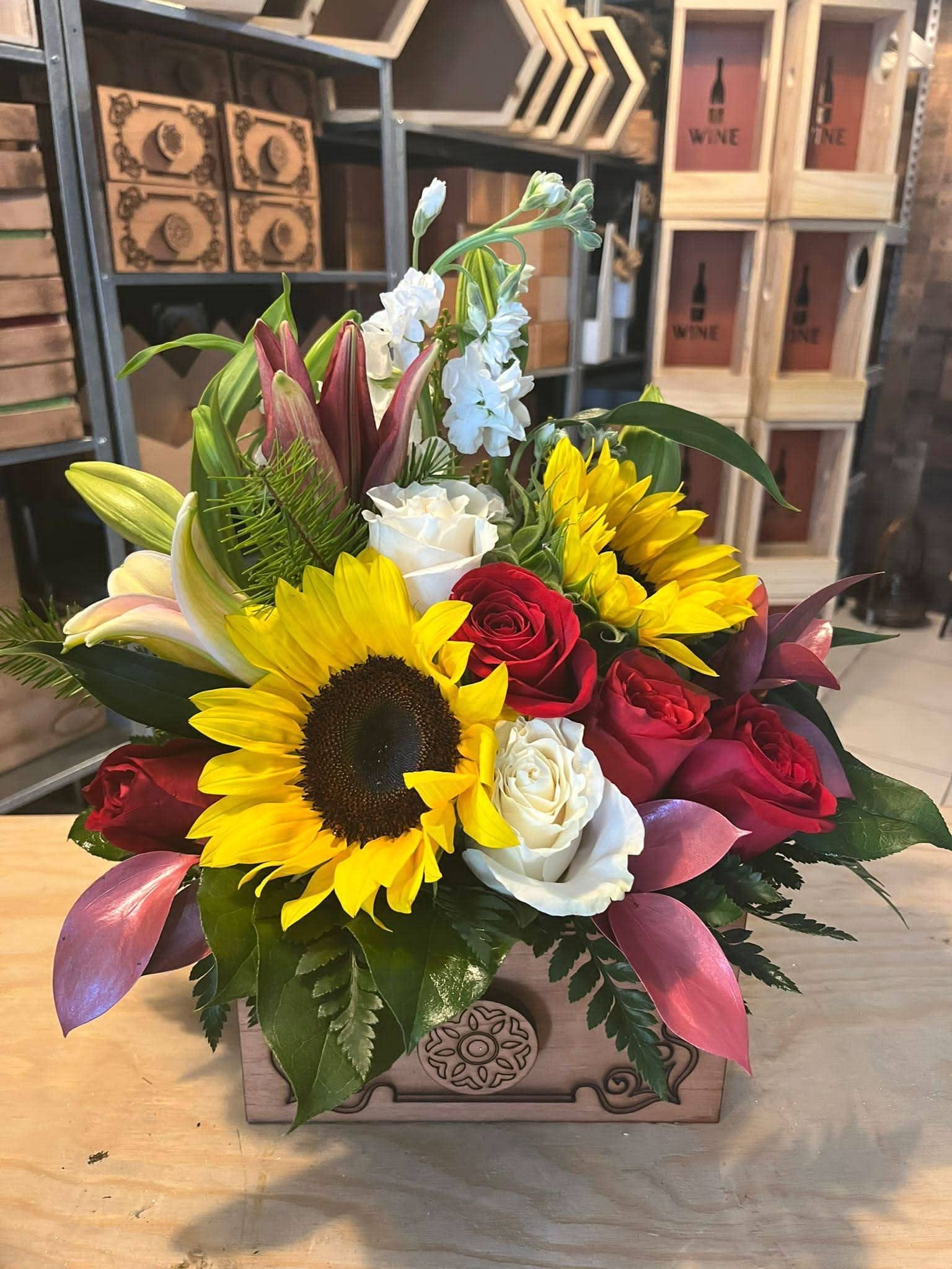 Arreglo floral con girasoles, rosas rojas, lirios y flores blancas en caja de madera decorativa.