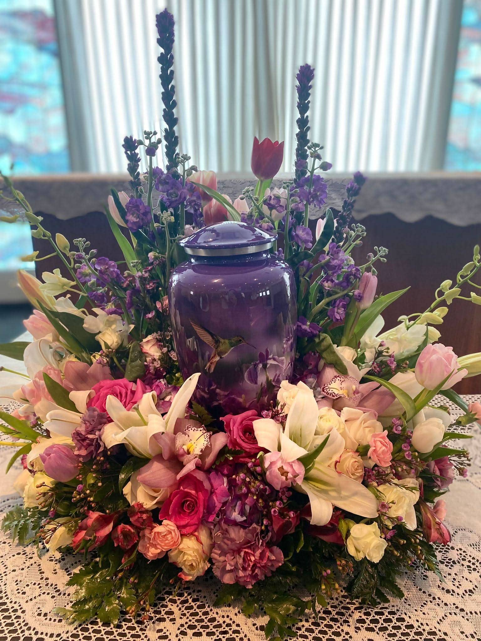 Porta cenizas morado rodeado de arreglo floral con lirios, rosas y tulipanes, decoración funeraria elegante.