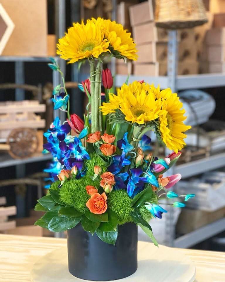 Arreglo floral colorido con girasoles, rosas naranjas y flores azules en base negra.