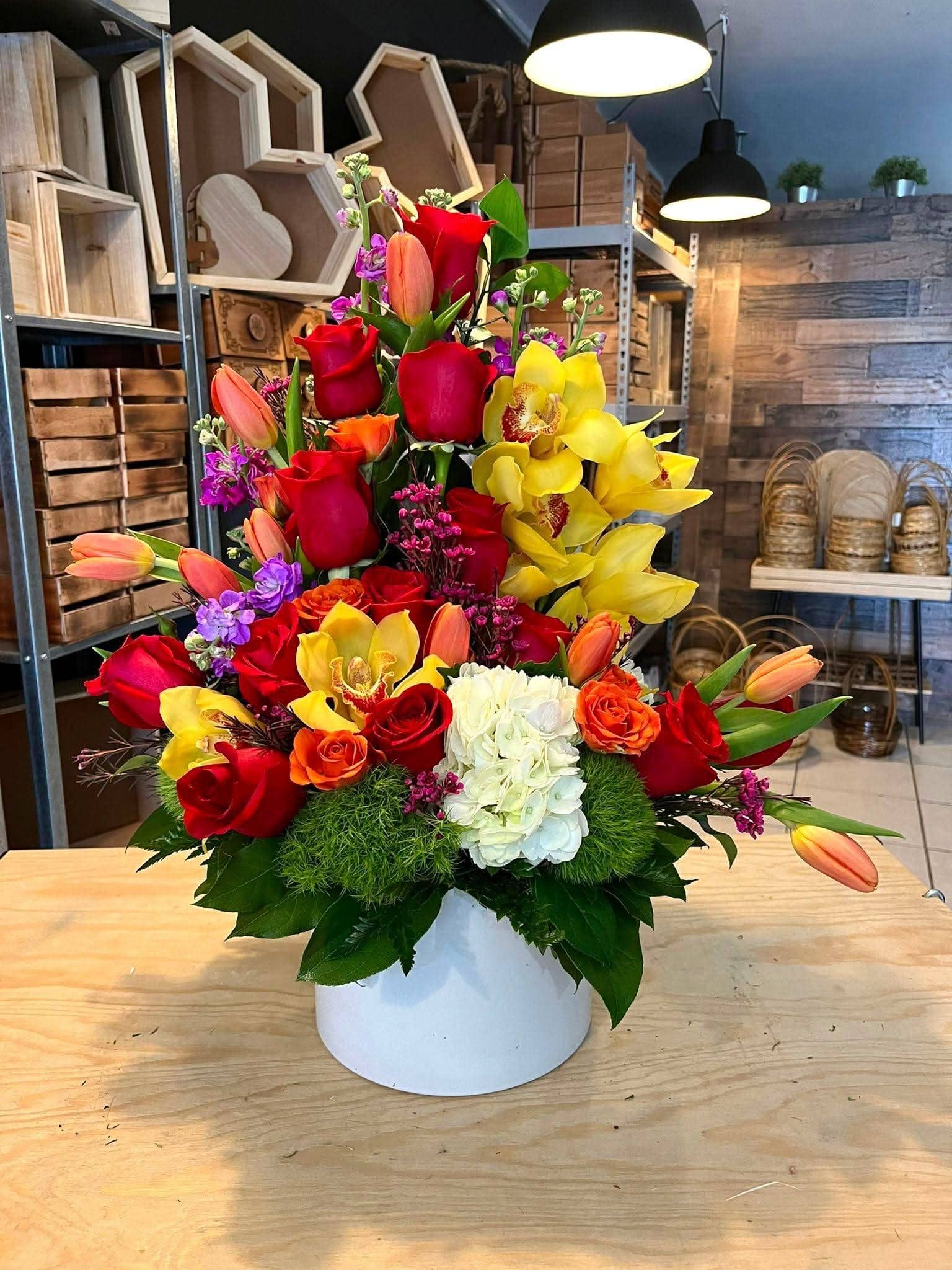 Arreglo floral colorido con rosas, tulipanes y orquídeas en base blanca sobre mesa de madera
